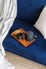 Ceramic teapot, cup of tea and croissant on breakfast wooden tray on couch
