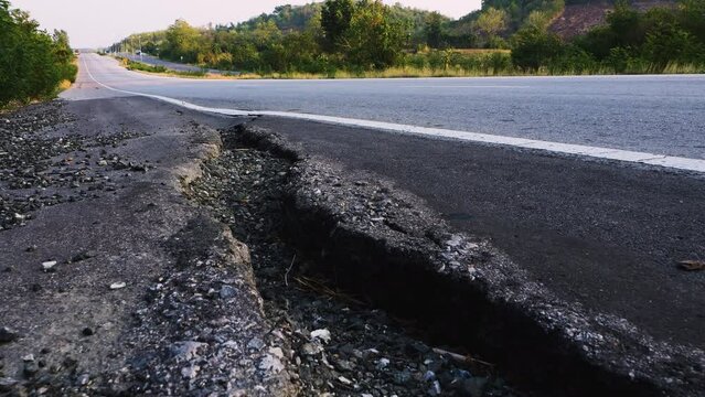 slow motion, The damaged road shoulder is cracked and the asphalt is broken, exposing the gravel beneath. Stones and debris are scattered across the area, vehicles continue to pass by.