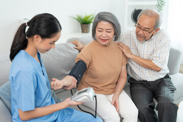 Asian caregiver doctor using digital pressure gauge measuring blood pressure of elderly woman patient Young female nurse in uniform gives consultation during home visit.
