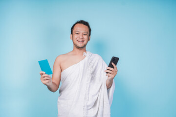 Happy young Asian Muslim man wearing ihram holding passport and smartphone while lifting head up...