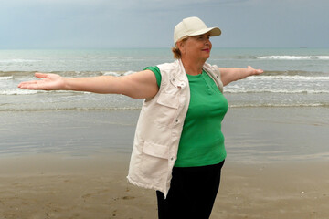 A plus size senior woman does exercises in the open air at beach against sky. Older female doing sport to keep fit. Concept of healthy living in the elderly.