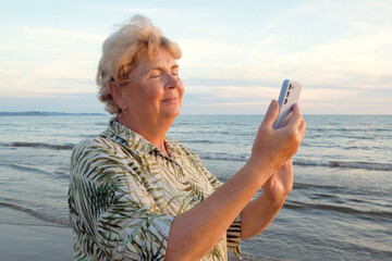 Senior woman staying on the sea beach and speaking with mobile phone in sunset rays. Mature woman smiles holding cellphone in hand. Concept of roaming, wifi, vacation, freedom and happy retirement.