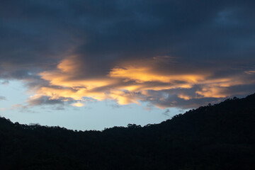Landscape of thick clouds, orange light hitting the blue sky, silhouettes of trees, forests, and mountains. Area for text in the middle of the image