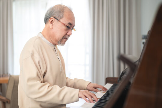 Happy relaxation Asian senior retirement man with eyeglasses learn to play piano at home, elderly old man playing music on piano in living room, lifestyle life after retirement, Close up hands