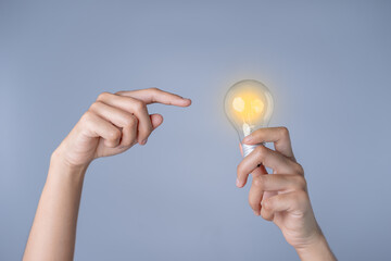 A young woman's hand points to a bright light bulb, a new innovation in science and technology. Renewable energy creative inspiration lies in a gray studio background.