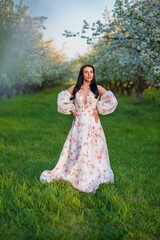 Fototapeta premium Full-length portrait of a beautiful brunette caucasian woman standing in blossoming apple garden in white floral dress on a fresh green grass