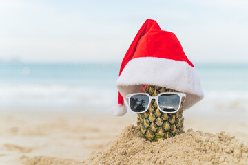 Santa Claus pineapple wears stylish sunglasses on the sand against the sea. wearing a christmas hat...