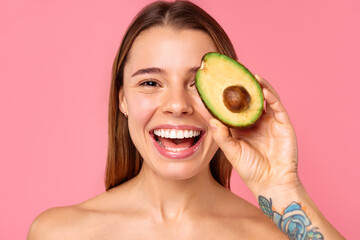 A woman joyfully holds half an avocado against her face, smiling, on a pink background