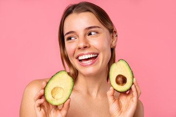 A woman joyfully holds half an avocado against her face, smiling, on a pink background