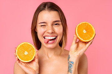 A woman is cheerfully holding orange halves for skin care, set against a pink background