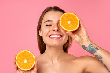 A woman is cheerfully holding orange halves for skin care, set against a pink background