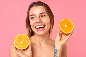 A woman is cheerfully holding orange halves for skin care, set against a pink background