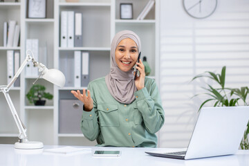 Confident businesswoman wearing a hijab talking on phone, sitting at desk with laptop in bright, modern office environment.