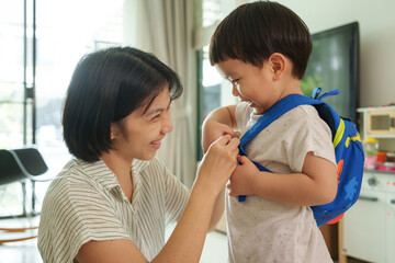 Smiling Asian mother adjusts blue backpack on happy young son. Child laughs while mother ensures...