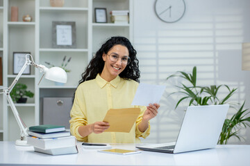 Smiling woman holding an envelope and letter while standing in a bright home office. Happy moment...