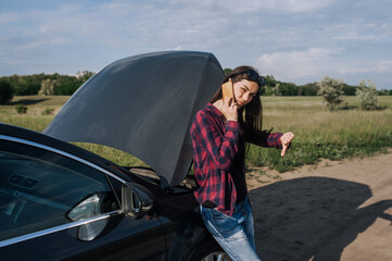 A young lonely brunette woman driver stands near a broken black car with an open hood on the side...