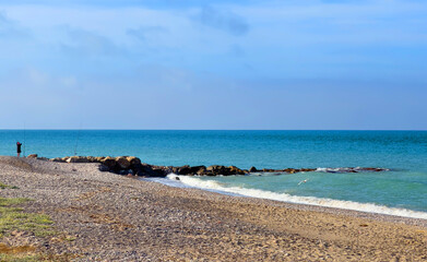 Empty stone beach. Breakwater with waves in sea. Fisherman catches fish on coast near sea. Catching fish in sea from shore. Fishing by sea. Beach with waves.