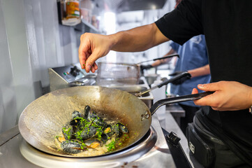 Close-up of a chef’s hands seasoning mussels in a pan on a stove, capturing the action and details of cooking