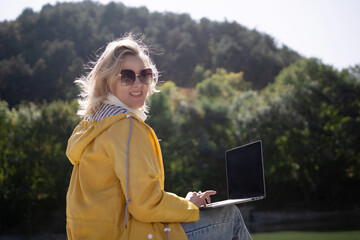 A woman in a yellow jacket is sitting on the grass with a laptop in front of her