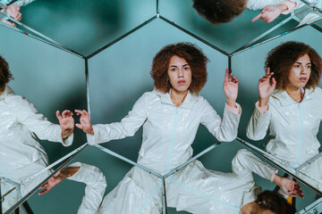 Young woman in white clothes posing in hexagon mirrors in front of grey wall with kaleidoscope...
