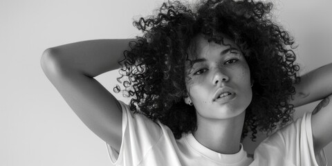 A woman with curly hair is posing for a photo. She is wearing a white shirt and has a tattoo on her arm. The image has a simple and elegant feel to it, with the woman's natural beauty