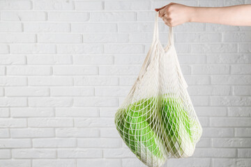 Woman holding string bag with fresh Chinese cabbages near white brick wall, closeup. Space for text