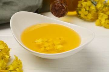 Rapeseed oil in gravy boat and beautiful yellow flowers on white wooden table, closeup