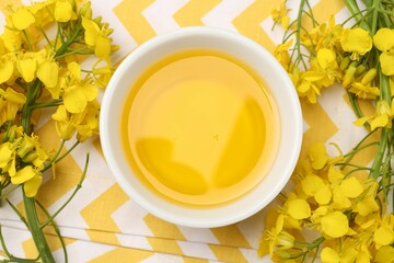 Rapeseed oil in bowl and beautiful yellow flowers on table, top view
