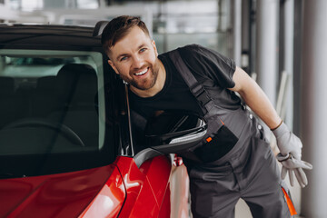 Bearded smiling auto mechanic opening the hood of a red car in a car repair shop