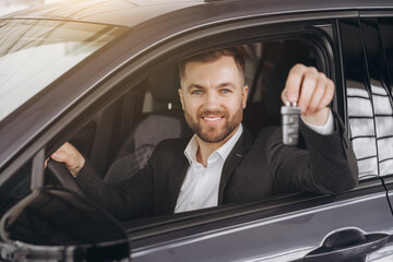 Cheerful bearded man holding key from auto, buying brand new car in showroom. Happy smiling guy buying new automobile, sitting inside car and showing automatic key at camera