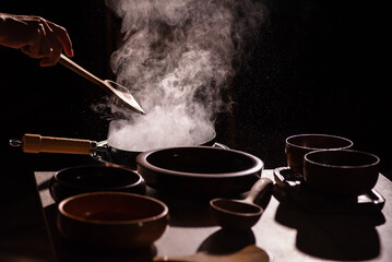 The cook was using a spatula to fry food in a pan of hot oil. There was a lot of smoke rising from the pan. There are wooden cups and plates placed on the table in a retro style.