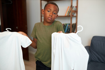 Puzzled african american male child shrugging shoulders holding two white t-shirts, trying to make...