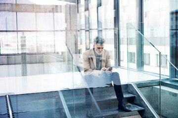 Handsome businessman sitting on stairs in modern office hallway, scrolling on smartphone.