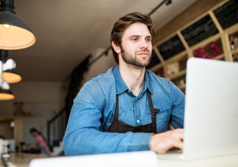Man in small business, working in his bistro, writing email to supplier. Young male business owner.