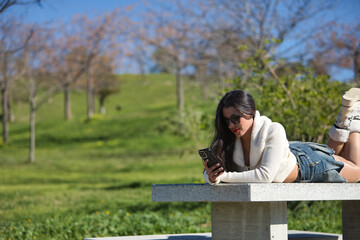 Fototapeta premium Young, beautiful, brunette woman in a white sweater, denim miniskirt and sunglasses, checking social media on her smartphone lying on a park bench on a sunny day. Beauty, fashion, trend concept.