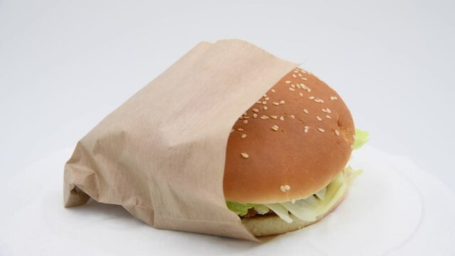 Close-up view of chickenburger with letuce salad in paper bag spinning against gray background. Bun covered with sesame seeds. Soft focus. Real time video. Fast food restaurant. Unhealthy eating theme