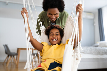 Playful African American little girl laughs as her mother pushes her on the swing in their bedroom.