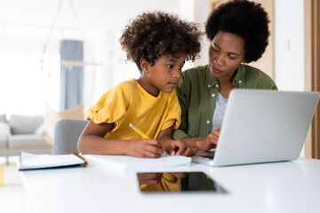 Attentive African American mother supervises her young daughter while she using laptop for homework.