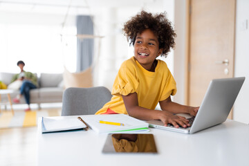 Smiling African American girl sitting at the table and using laptop. Her mother is sitting on the...