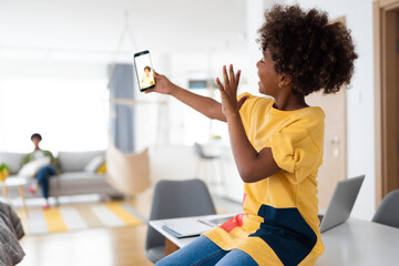 Cute African American young girl using mobile phone talking on a video call with friend while her mother is sitting on the sofa in the background. - Powered by Adobe