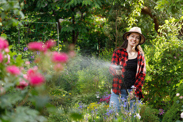 smiling young woman gardener watering flowers and plants in garden with hose