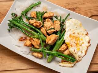 Asian dish with fried chicken, broccolini, fried egg and wide noodles served on a white plate on a wooden board.