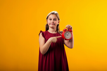 Smiling kid girl holding an alarm clock in hand and pointing finger at it. The concept of education, school, deadlines, time to study