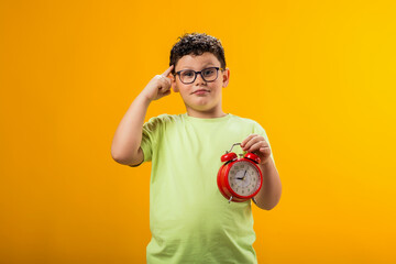Thoughtful child boy holding alarm clock over yellow background and scratching head. Time...