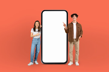 A man and woman are standing in front of a blank screen, both pointing towards it, against a...