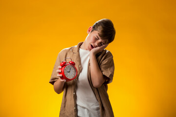 Boy holding alarm clock and showing sleeping gesture over yellow background. Time management concept