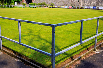 Fototapeta premium Lush green lawn bowling field with railings, in bright sunlight, surrounded by walls and trees
