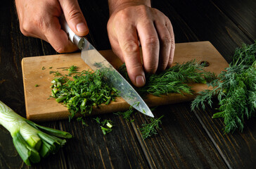 Chef hands use a knife to cut fresh dill on a cutting board for preparing a vegetarian dish....