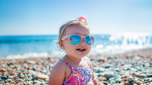 beb&eacute; femenino tierno y feliz en la arena de la playa con gafas de sol, mirando de frente a la camara 
