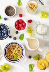 Top view different ingredients for cooking a healthy breakfast setup with granola, assorted fresh fruits, chia seeds and yogurt on a white marble background